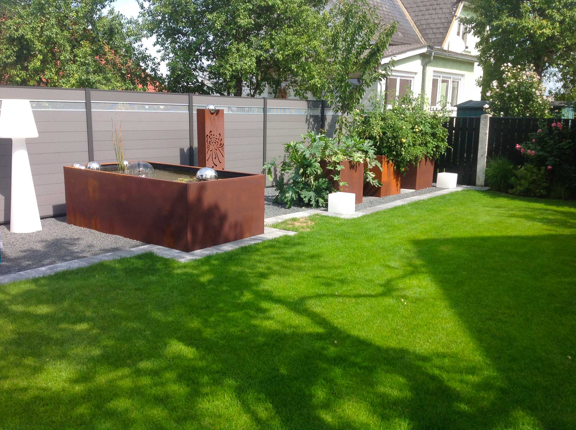 Moderner Garten mit rostfarbenen Pflanzgefäßen und rechteckigem Wasserspiel vor grauem Zaun Garten mit grünem Rasen, braunen Pflanzkübeln, Wasserspiel und grauem Sichtschutzzaun bei sonnigem Wetter