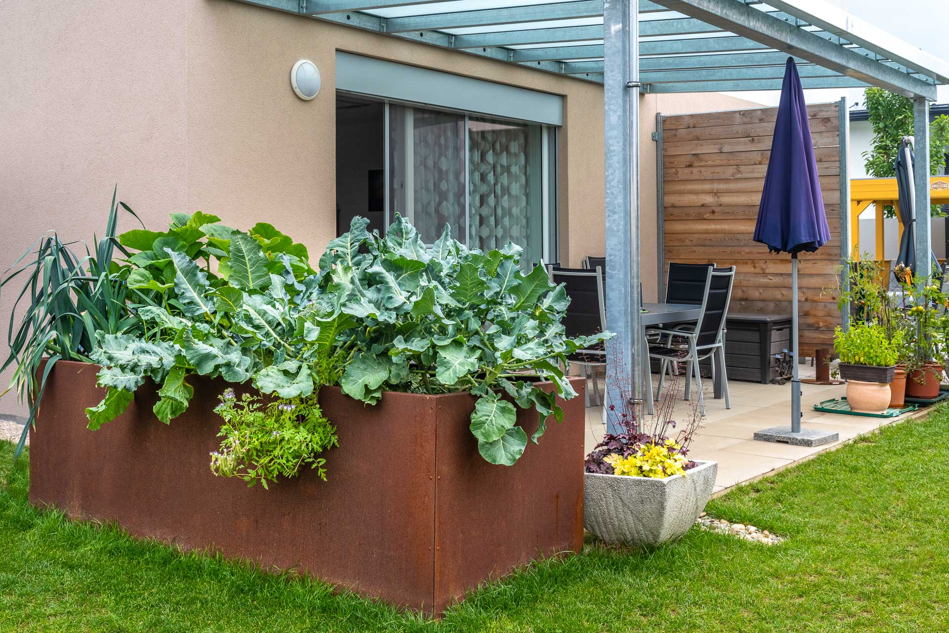 Moderner Hochbeet-Garten aus Stahl vor Terrasse mit Überdachung und Sitzbereich Großes Roststahl-Hochbeet mit verschiedenen Pflanzen vor einer Terrasse mit Glasüberdachung, Gartentisch mit Stühlen und geschlossenen Sonnenschirm