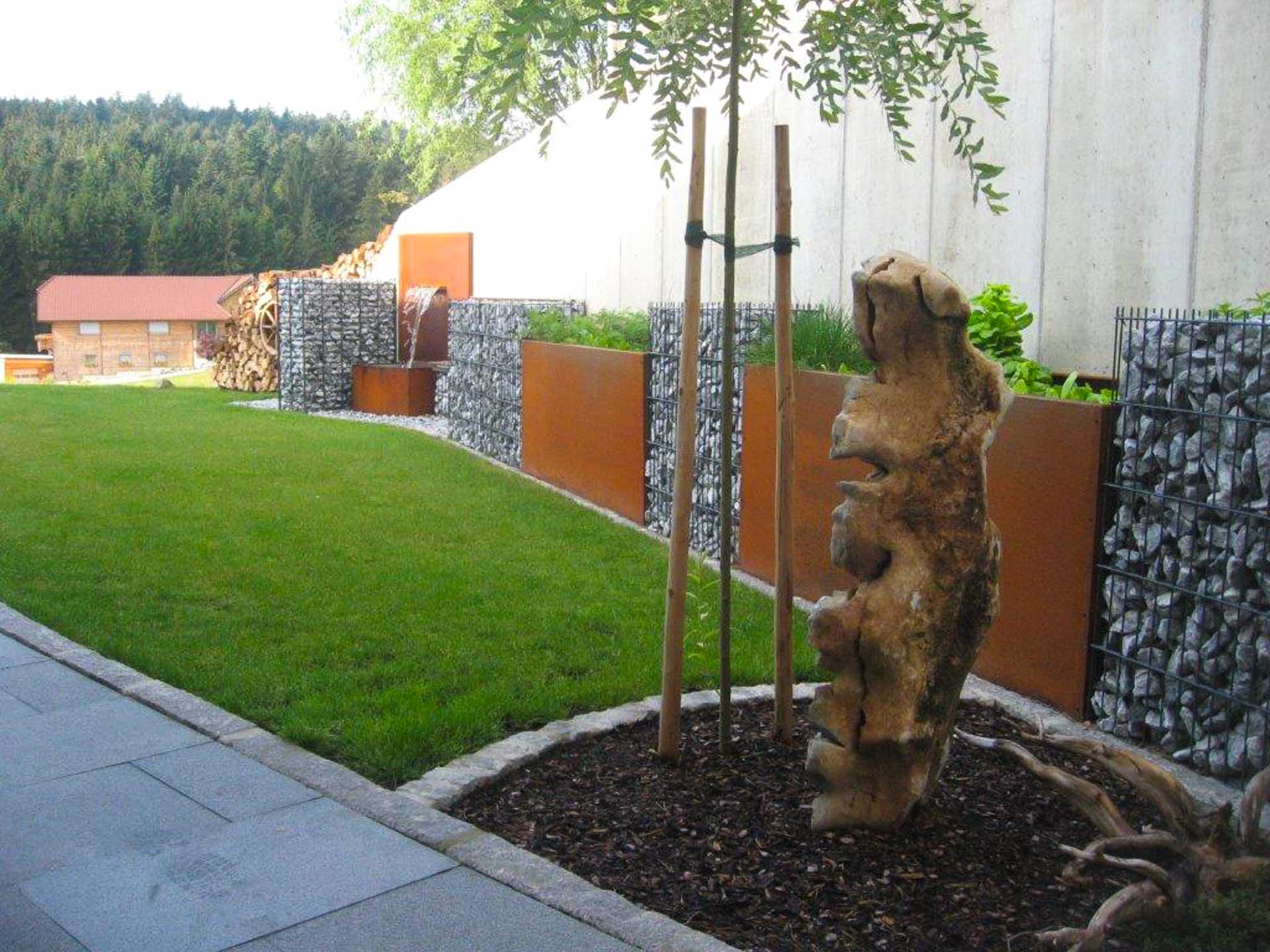 Garten mit grüner Rasenfläche, Gabionenwänden aus Steinkörben mit Cortenstahlelementen, Wasserbrunnen und dekorativem Baumstamm im Vordergrund