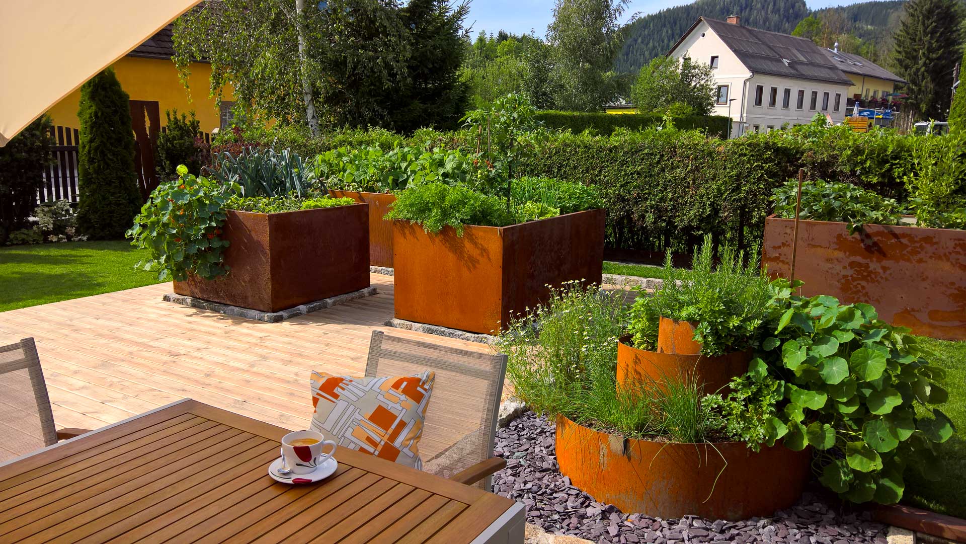 Garten mit mehreren rostbraunen Hochbeeten, bepflanzt mit frischen Kräutern und Gemüse, Holzterrasse mit Stühlen und Tisch, Tasse Kaffee auf dem Tisch