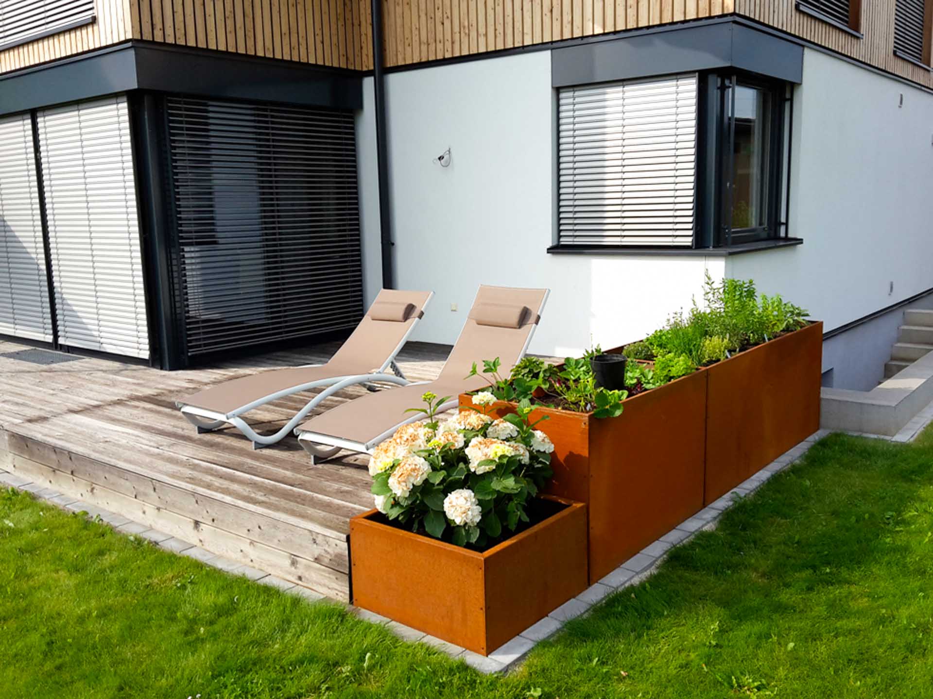 Moderne Terrasse mit zwei Sonnenliegen und bepflanzten Hochbeeten aus Cortenstahl neben einem weißen Gebäude Terrasse mit zwei beigen Sonnenliegen und Hochbeeten aus rostfarbenem Cortenstahl, bepflanzt mit Blumen und Kräutern vor einem modernen Haus mit weißen Wänden und Holzverkleidung
