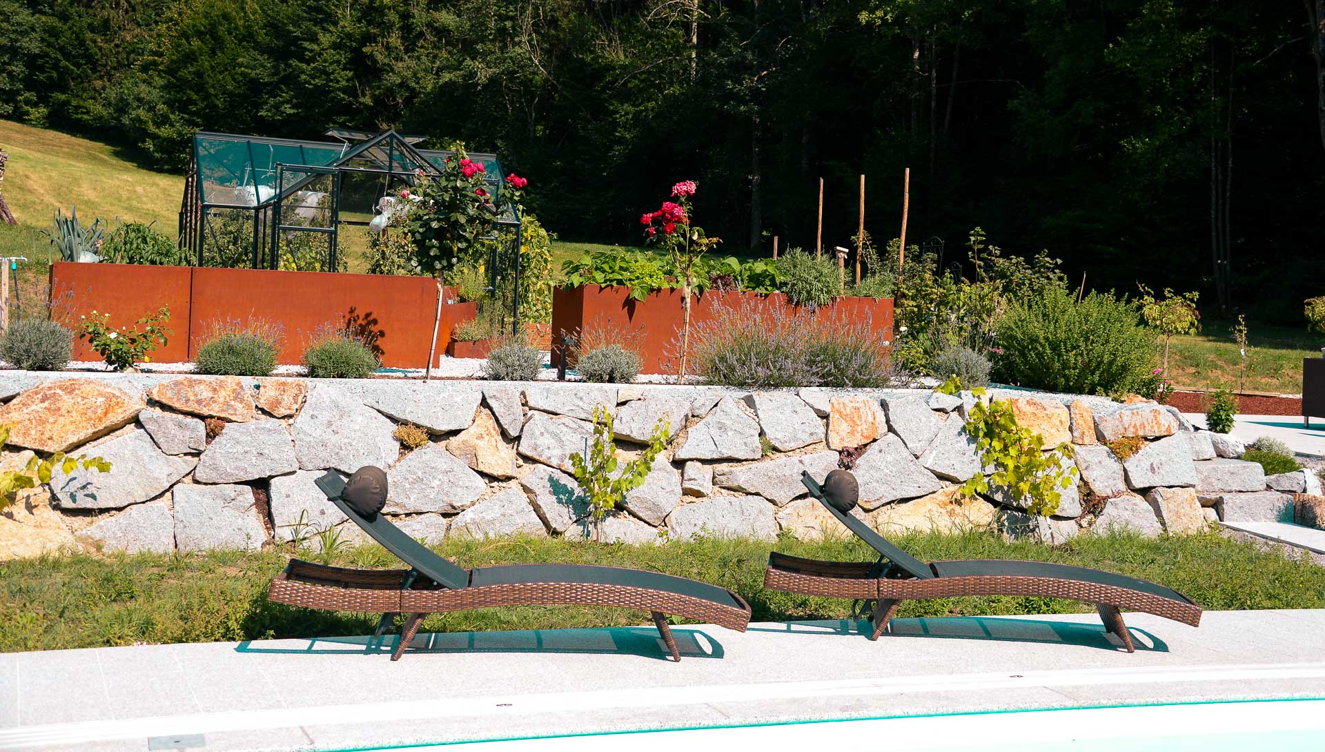 Zwei bequeme Rattan Liegestühle neben einem Pool mit Steinterrasse und bepflanzten Hochbeeten im Garten Zwei dunkle Rattan Liegestühle am Pool mit Steinterrasse und bepflanzten Hochbeeten im Hintergrund
